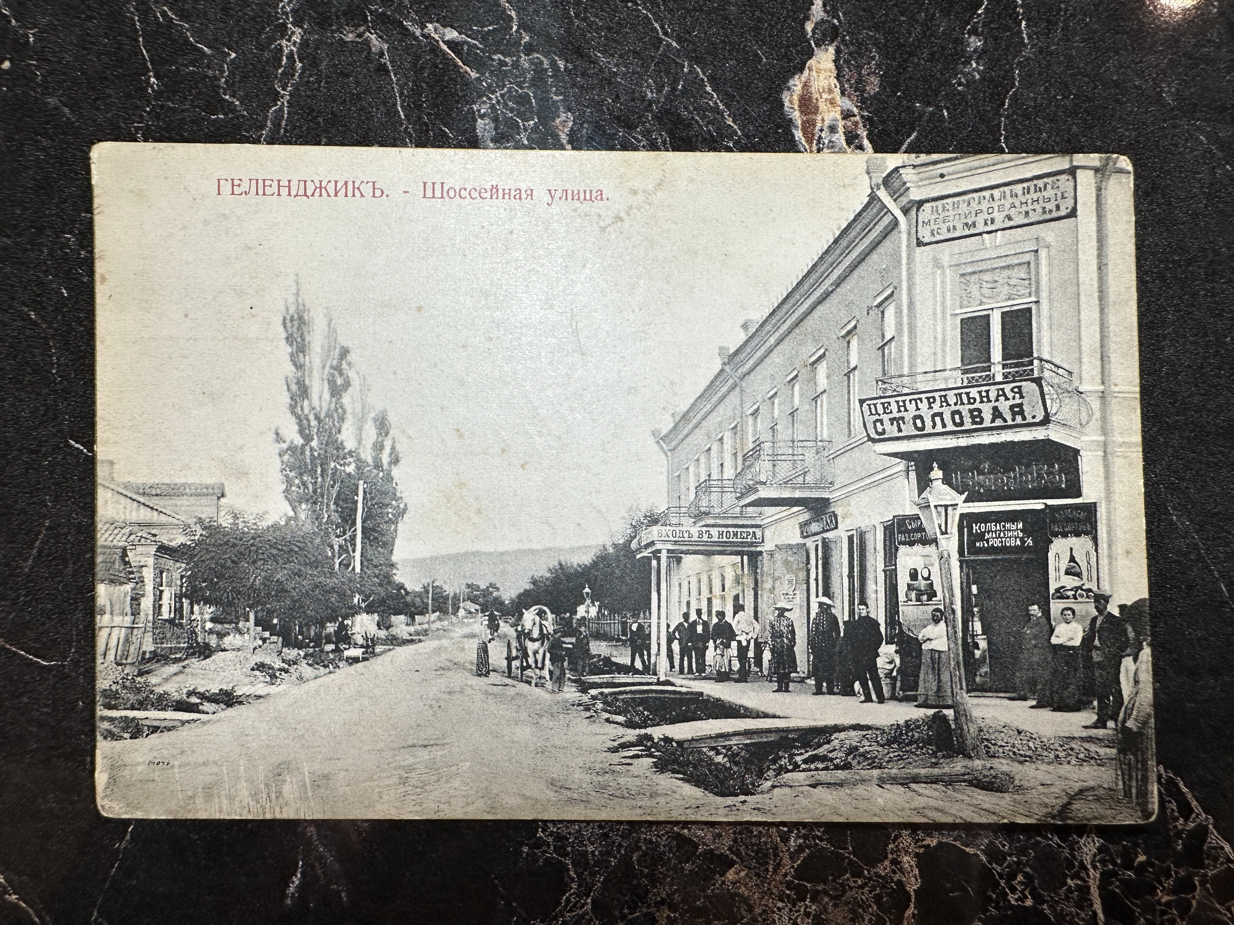 Черно-белая дореволюционная открытка с видом на Шоссейную улицу в Геленджике, изображающая здание Центральной столовой, вывески магазинов и людей.